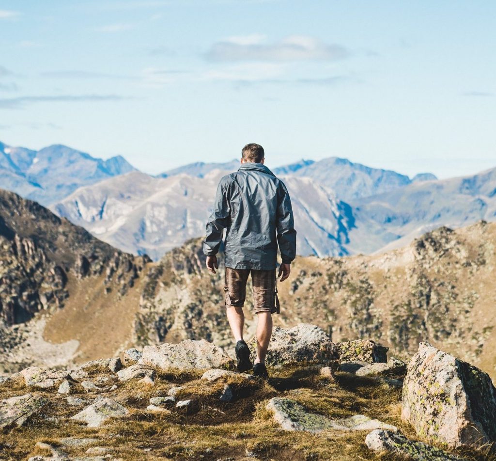 Wanderer auf Fels – ©StockSnap/Pixabay Ein Mann, von Hinten zu sehen, steht auf einem Fels und blickt auf das Gebirge unter ihm.