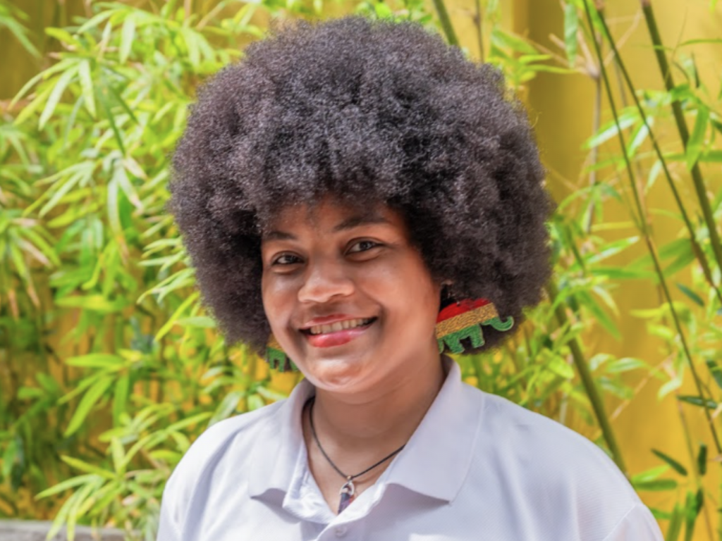 Hotel employee wearing a white polo shirt, standing in front of plants and looking directly at the camera.